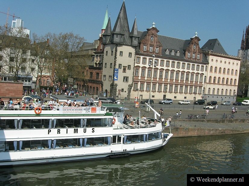 In de rivier de Main ligt een rondvaartboot vlakbij het plein Römerberg in de Altstadt. Het grote gebouw met de toren is het Historisches Museum. De voorzijde ligt aan het plein Römerberg.