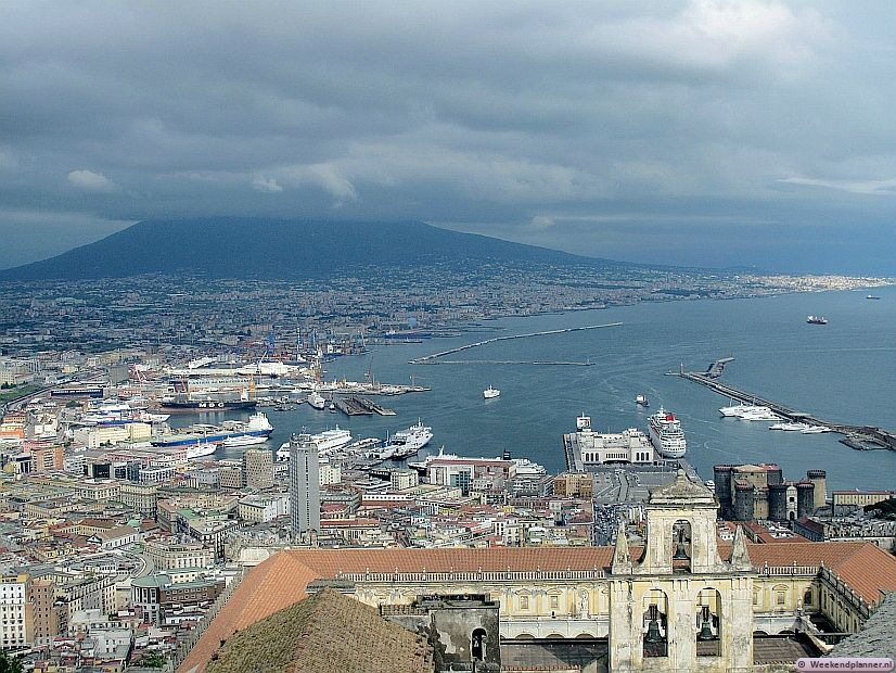 Dit is Napels met op de achtergrond de vulkaan Vesuvius. In de haven van Napels liggen cruiseschepen en er liggen veerboten naar eilanden als Capri en Ischia. Foto: Het uitzicht vanaf de muren van kasteel Sant'Elmo op Napels. Tips: De attracties van de Golf van Napels.